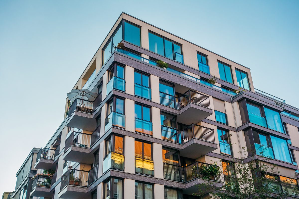 A multi-storey apartment building with glass balconies and large windows, seen from below against a blue sky